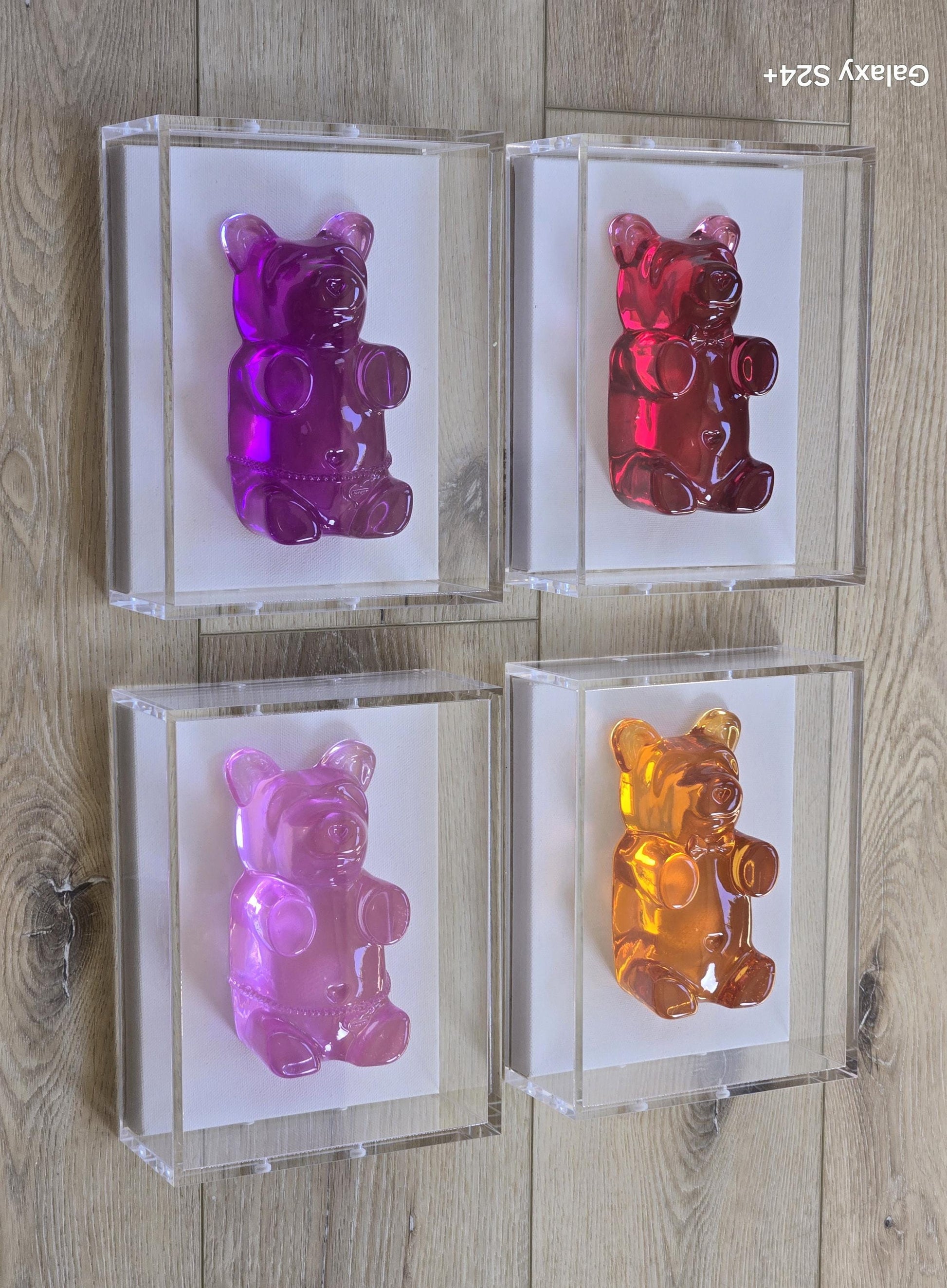 May include: Four framed gummy bear sculptures in shades of purple, red, pink, and orange. Each bear is encased in a clear acrylic box, mounted on a white background. The artwork is displayed against a light wood-grain surface.